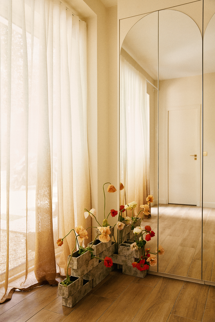 Une pièce avec de longues rideaux beiges, un grand miroir mural et un sol en bois. Devant le miroir, il y a un arrangement de fleurs en pot dans des blocs de béton.