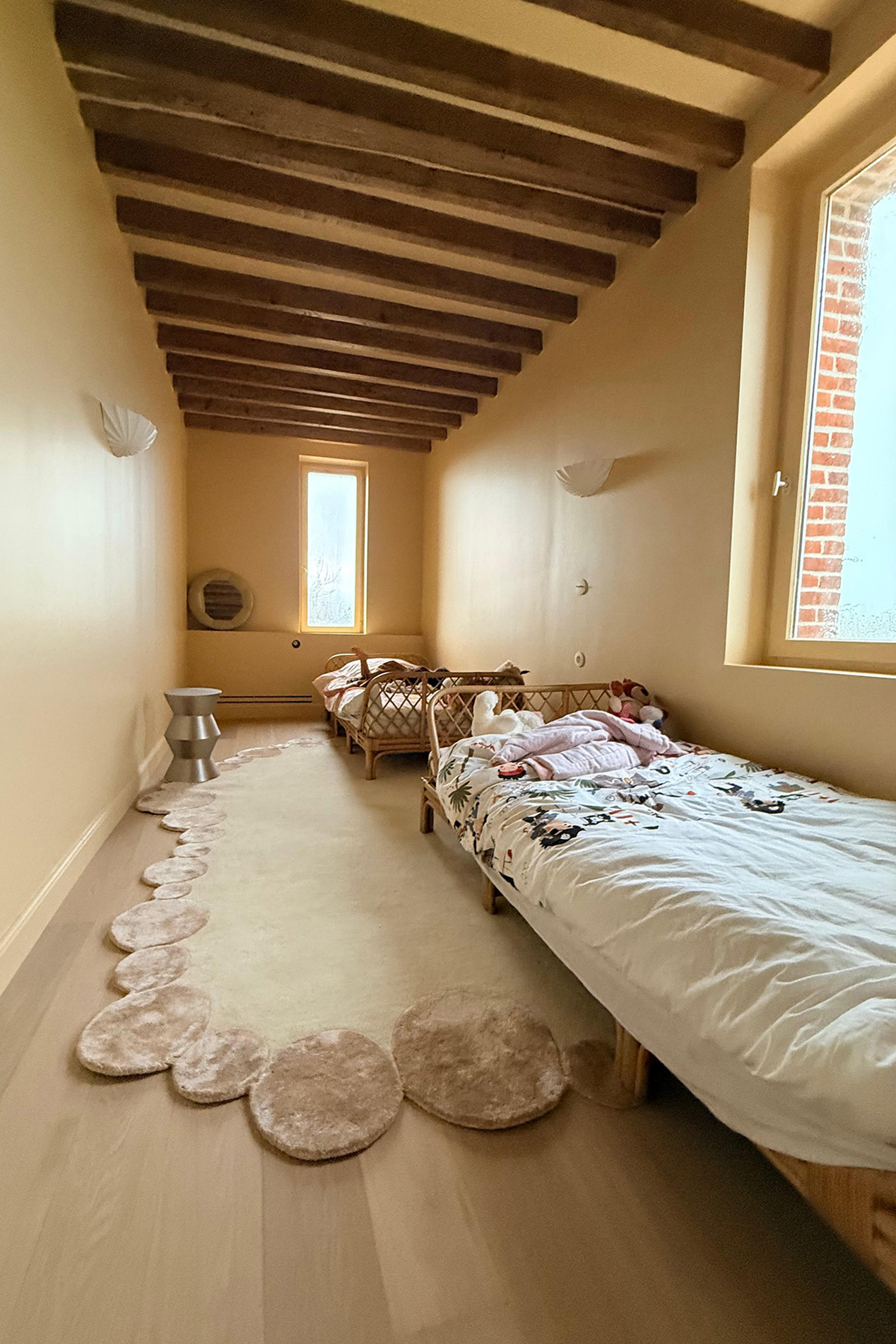 Chambre avec deux lits, un tapis beige avec des motifs de pétales autour, une fenêtre, des murs jaunes, et un plafond avec poutres en bois.
