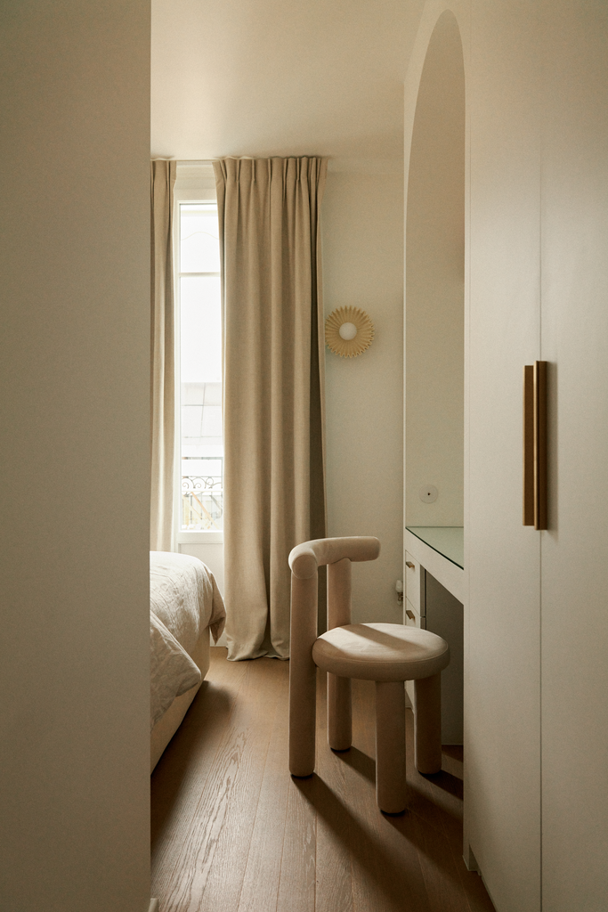 Une chambre élégante avec un lit, une fenêtre avec rideaux beiges, un miroir rond au mur, un bureau blanc avec une chaise beige et une armoire blanche.