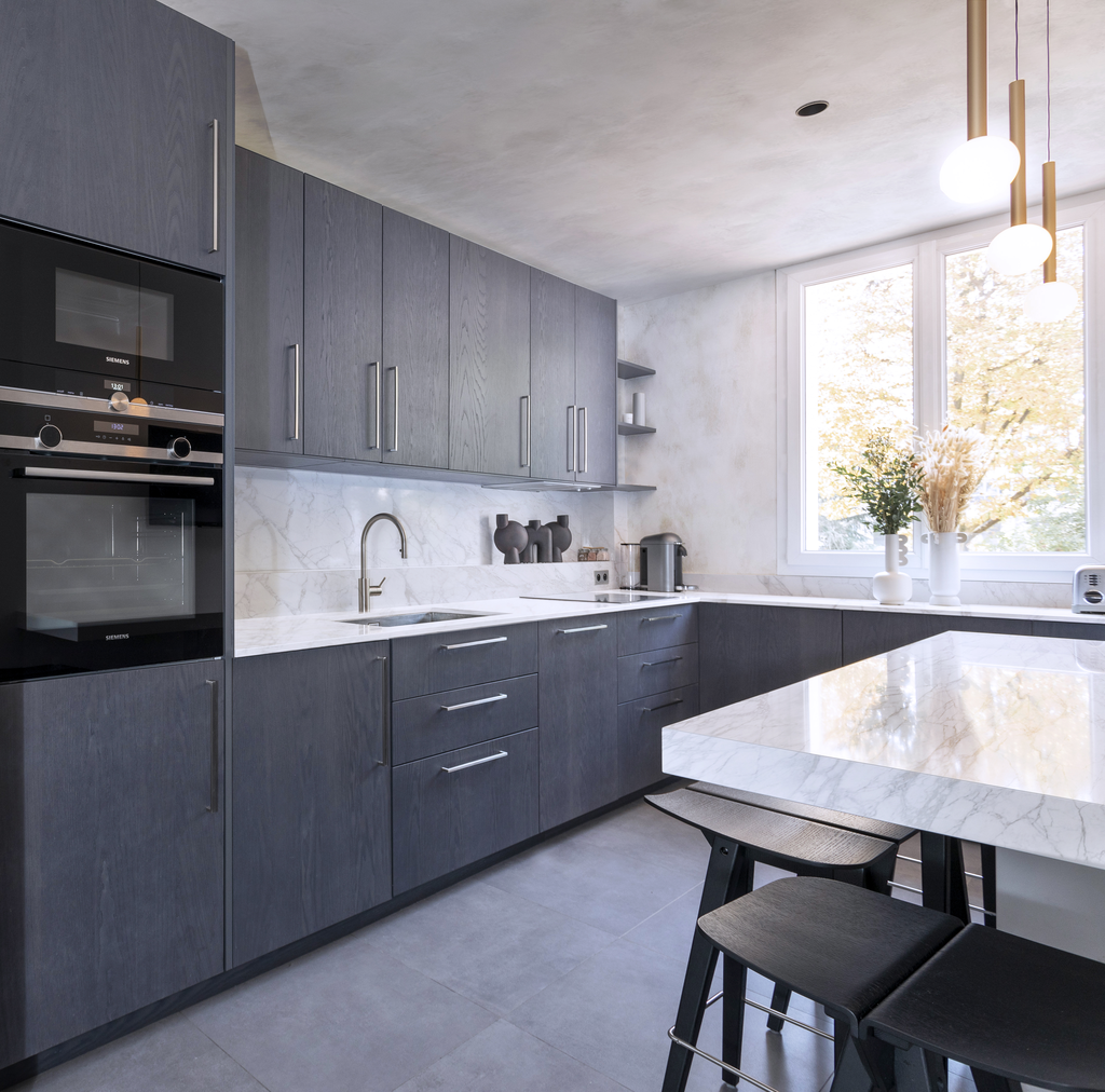 Cuisine moderne avec armoires en bois gris, comptoir en marbre blanc, fenêtres grandes laissant entrer la lumière naturelle, et décoration minimaliste avec des vases et des appareils électroménagers.