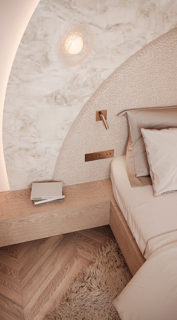 Vue partielle d'un lit avec une tête de lit en tissu beige, une table de nuit en bois clair avec deux livres empilés, un mur en pierre en arrière-plan, et un plafonnier décoratif