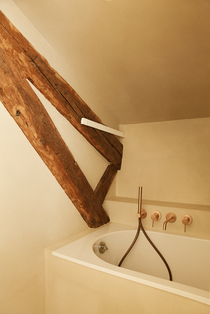 Une baignoire dans une pièce avec un plafond en pente, des poutres en bois apparentes, et des robinets muraux roses.