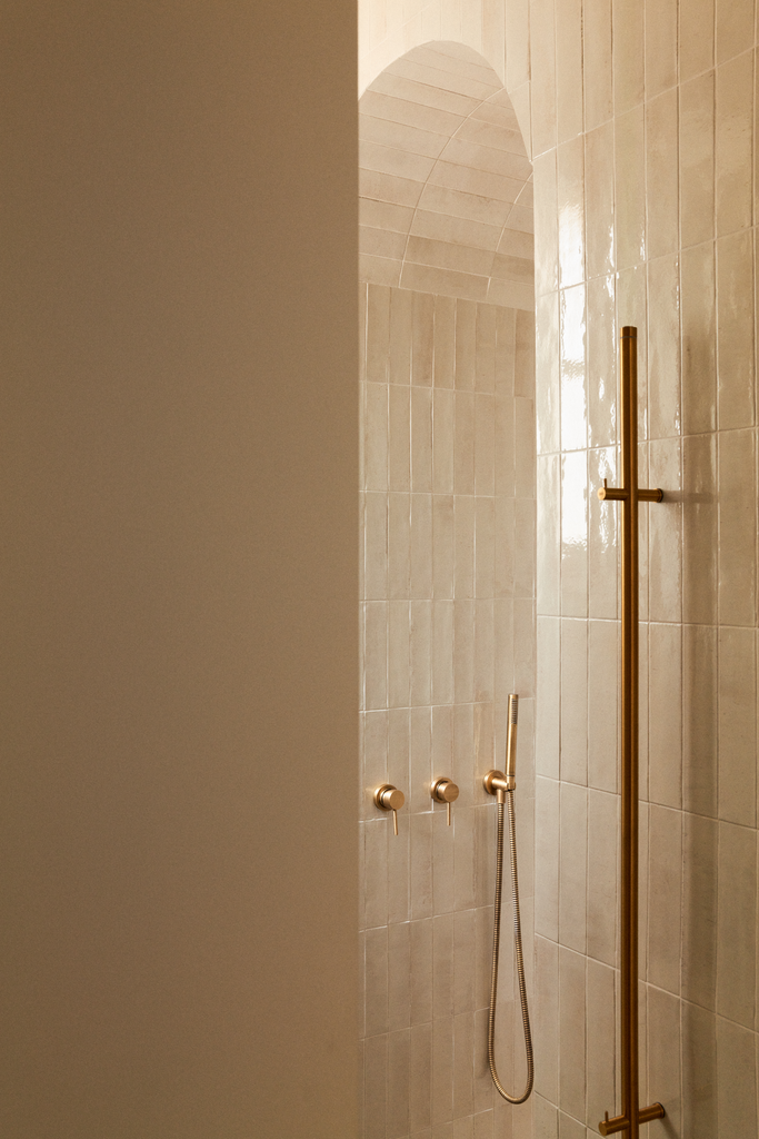 Vue d'une douche dans une salle de bain avec des carreaux beige, une barre de douche en métal doré, un pommeau de douche fixé au mur, et une arche en béton au plafond.