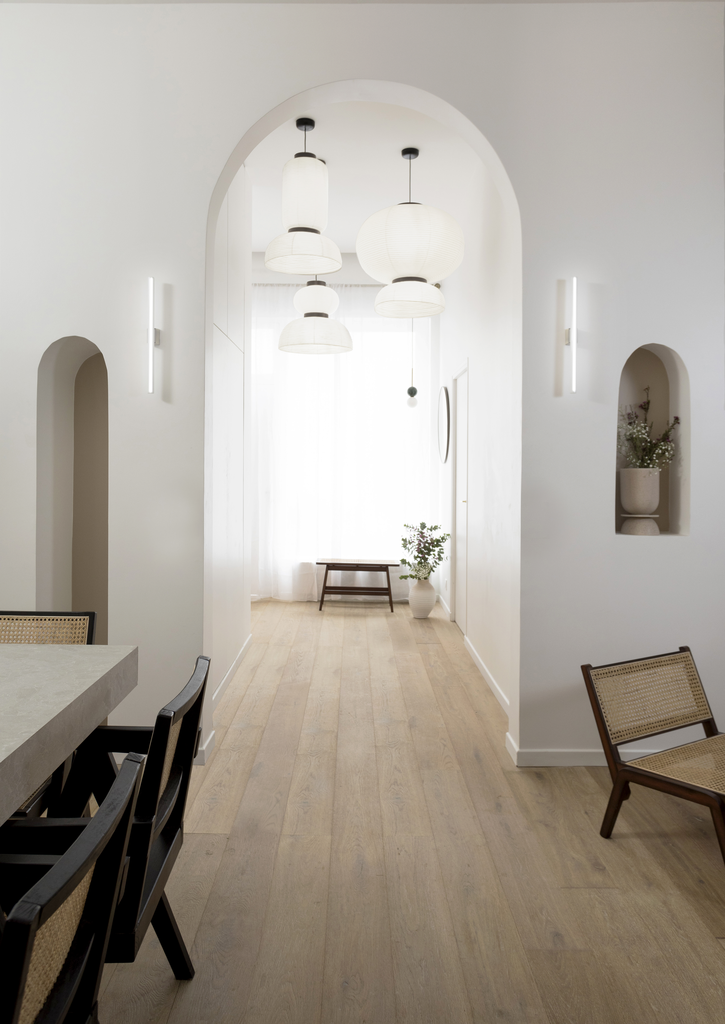 Intérieur lumineux avec un plafond arqué, des tapisseries suspendues, un sol en bois clair, des lampes en papier flottantes, une table avec des chaises en bois, une plante dans un vase blanc, et une niche intégrée avec des fleurs.