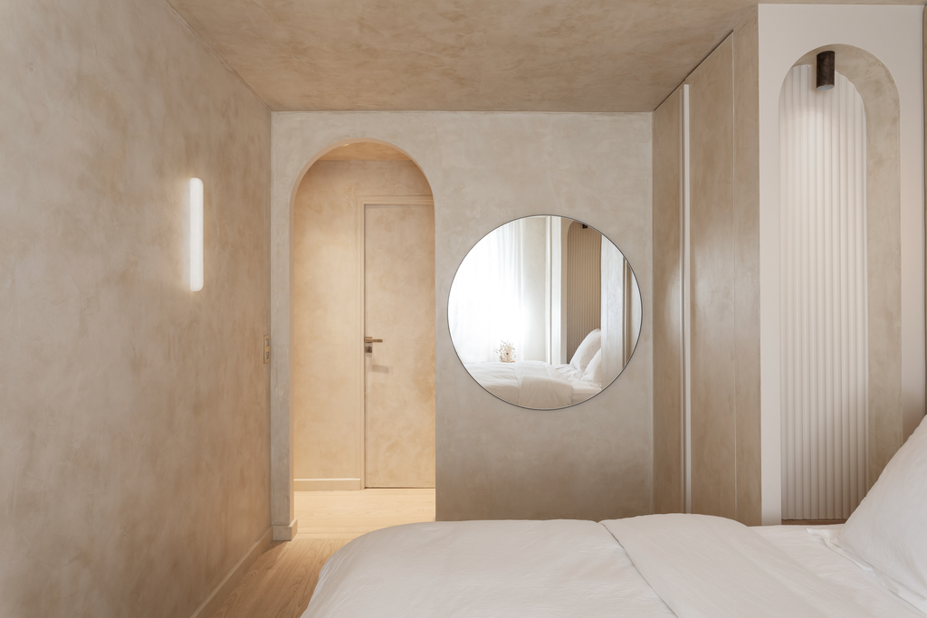 Chambre minimaliste avec murs beige, miroir rond, porte et fenêtre en arrière-plan, lit blanc.