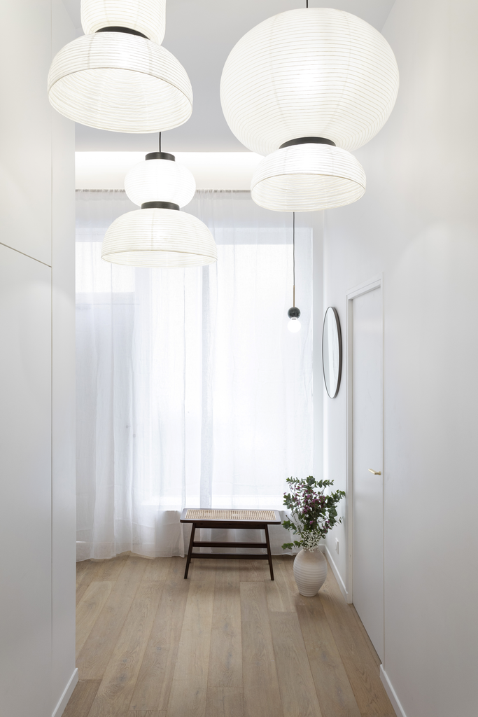 Un couloir lumineux avec des lampes en papier blanc suspendues au plafond, une fenêtre avec des rideaux blancs, un banc en bois et une grande plante dans un vase blanc.