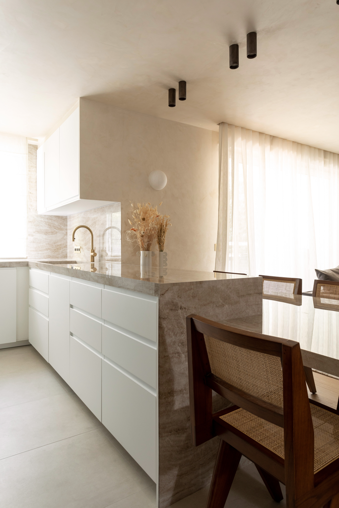 Cuisine moderne avec comptoir en marbre, luminaires noirs au plafond, voilage blanc laissant passer la lumière, chaises en bois autour d'une table, décoration avec des plantes séchées dans un vase blanc.