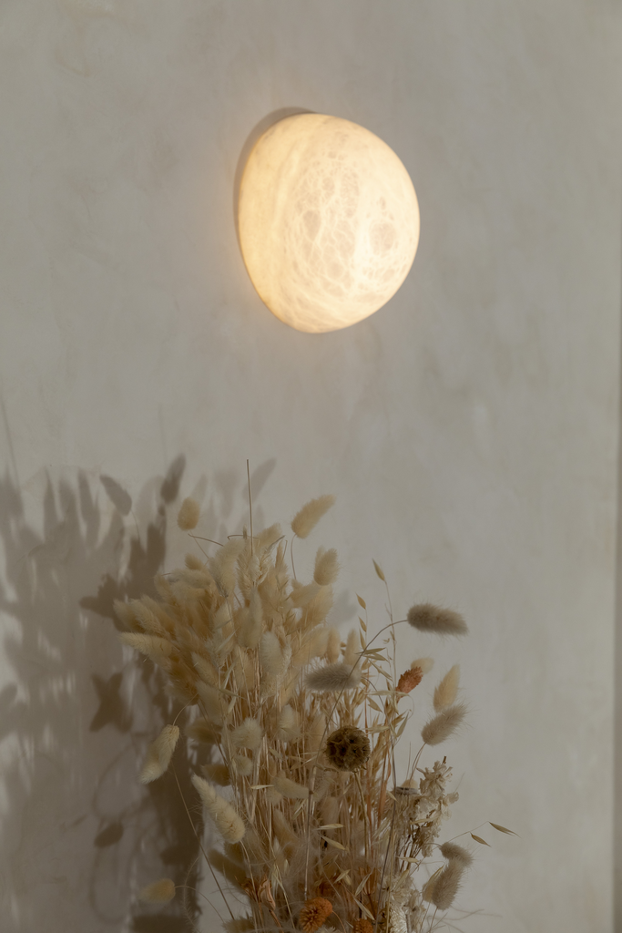 Une lampe murale en forme de lune illuminée, située au-dessus d'un bouquet de fleurs séchées en nuances neutres, avec un mur beige clair en toile de fond.
