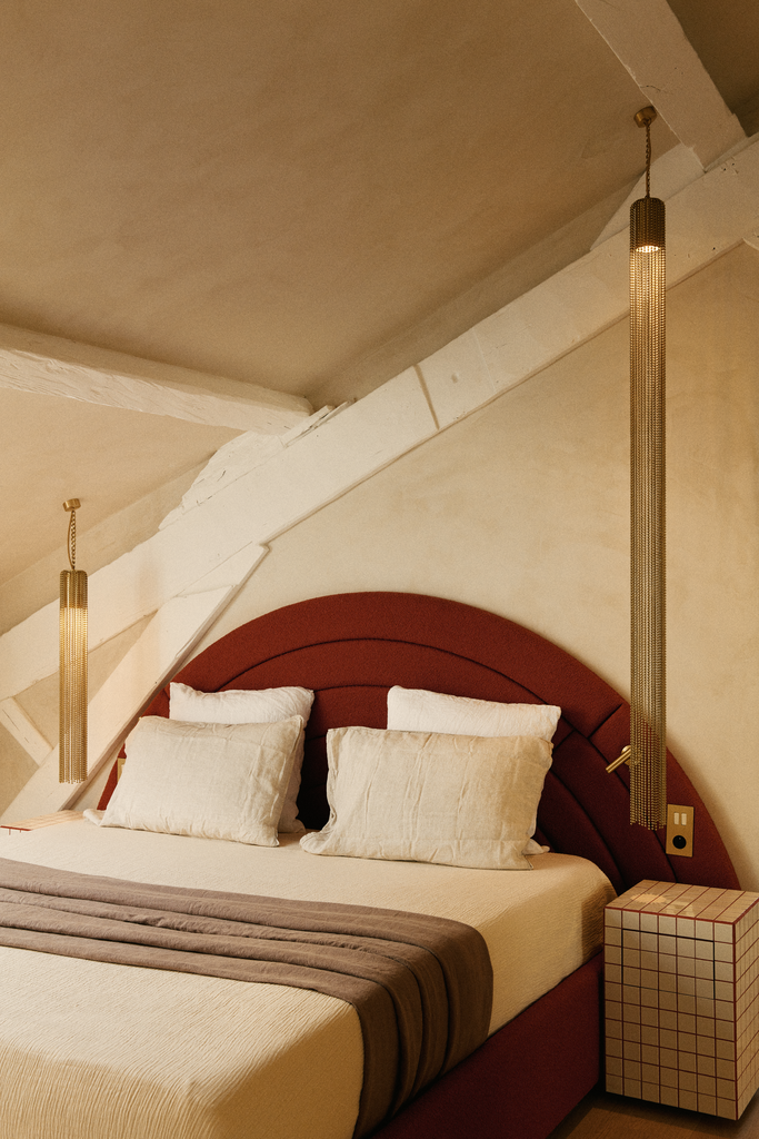 Un lit double avec une tête de lit rouge et plusieurs oreillers, dans une chambre avec un plafond en pente et deux lampes suspendues dorées.