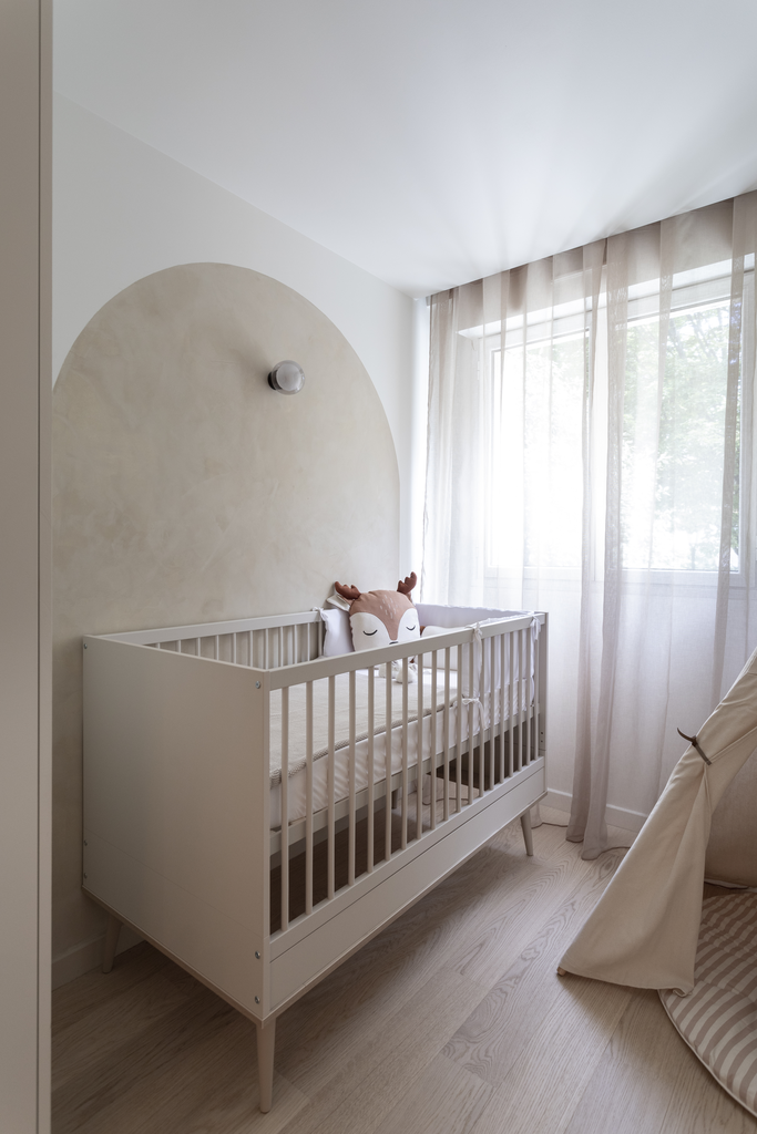 Une chambre d'enfant avec un berceau blanc, un coussin en forme de visage de renard, et une grande fenêtre avec des rideaux légers laissant entrer la lumière naturelle.