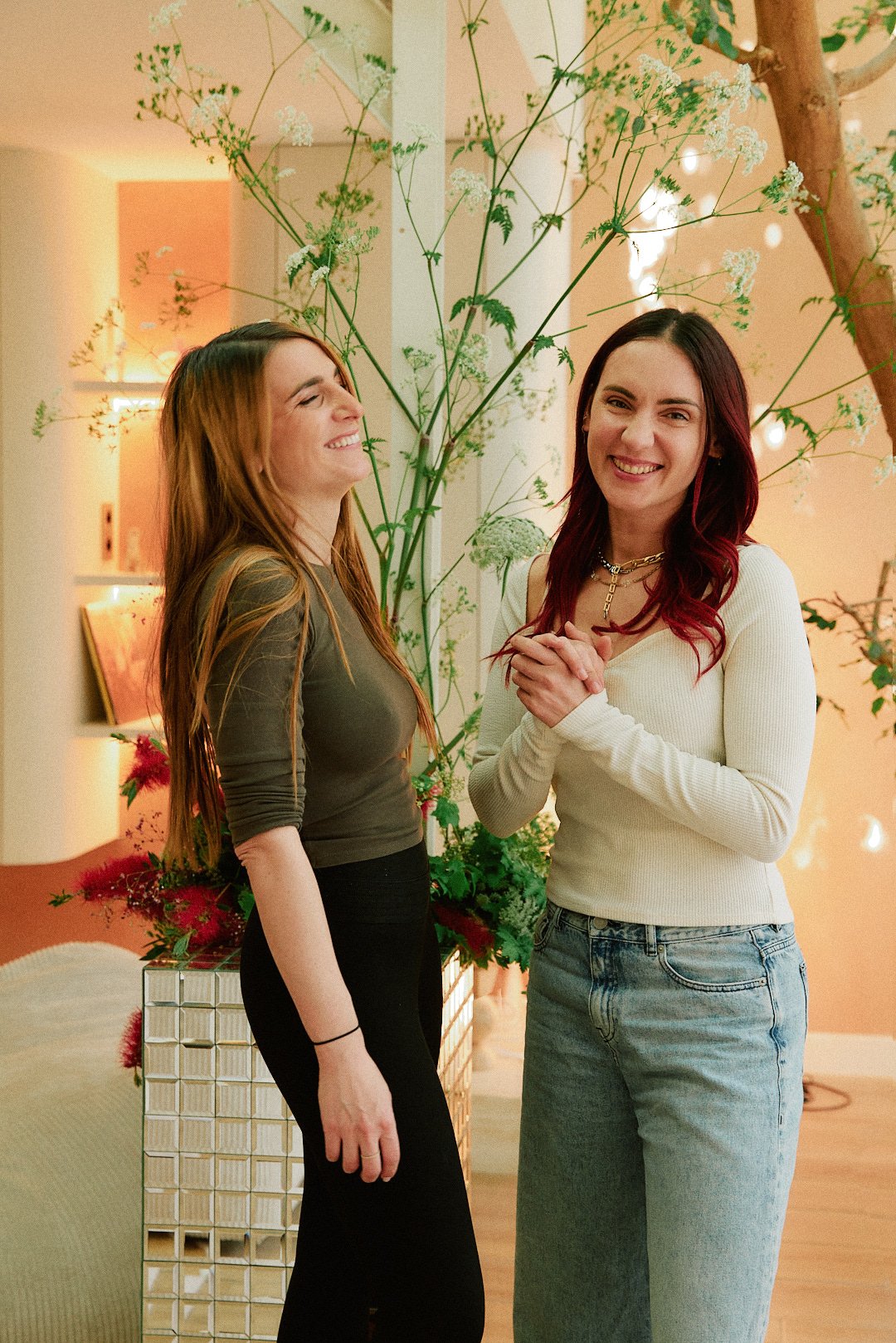 Deux femmes souriantes debout dans un intérieur décoré avec des plantes vertes et des lumières douces.
