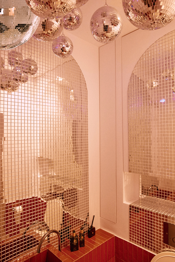 Pièce avec de nombreux miroirs disco suspendus au plafond et un mur en mosaïque de carreaux brillants, avec un lavabo et des produits de beauté sur le comptoir.