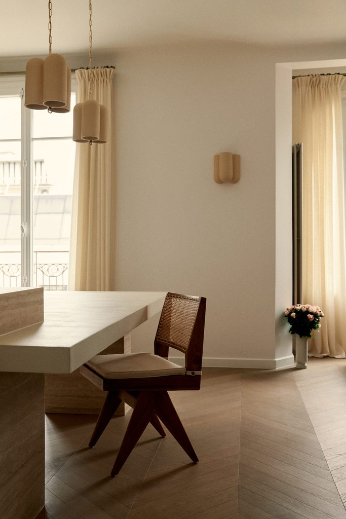 Intérieur lumineux avec un mur blanc, fenêtre avec rideaux beiges, une table blanche, une chaise avec cadre en bois et assise en tissu beige, et une plante en pot avec des fleurs.