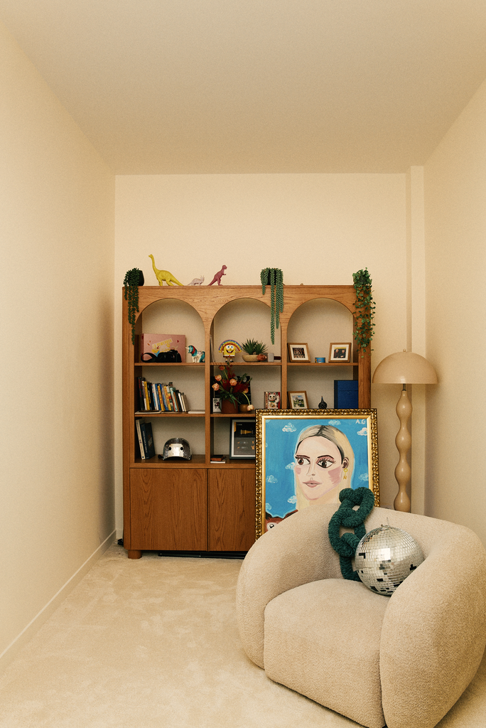 Salon avec un meuble en bois, plusieurs livres, décorations, et un tableau de portrait encadré. Un fauteuil beige avec un miroir disco et une chaîne en peluche verte. Lampe à pied et quelques figurines et plantes sur le meuble.