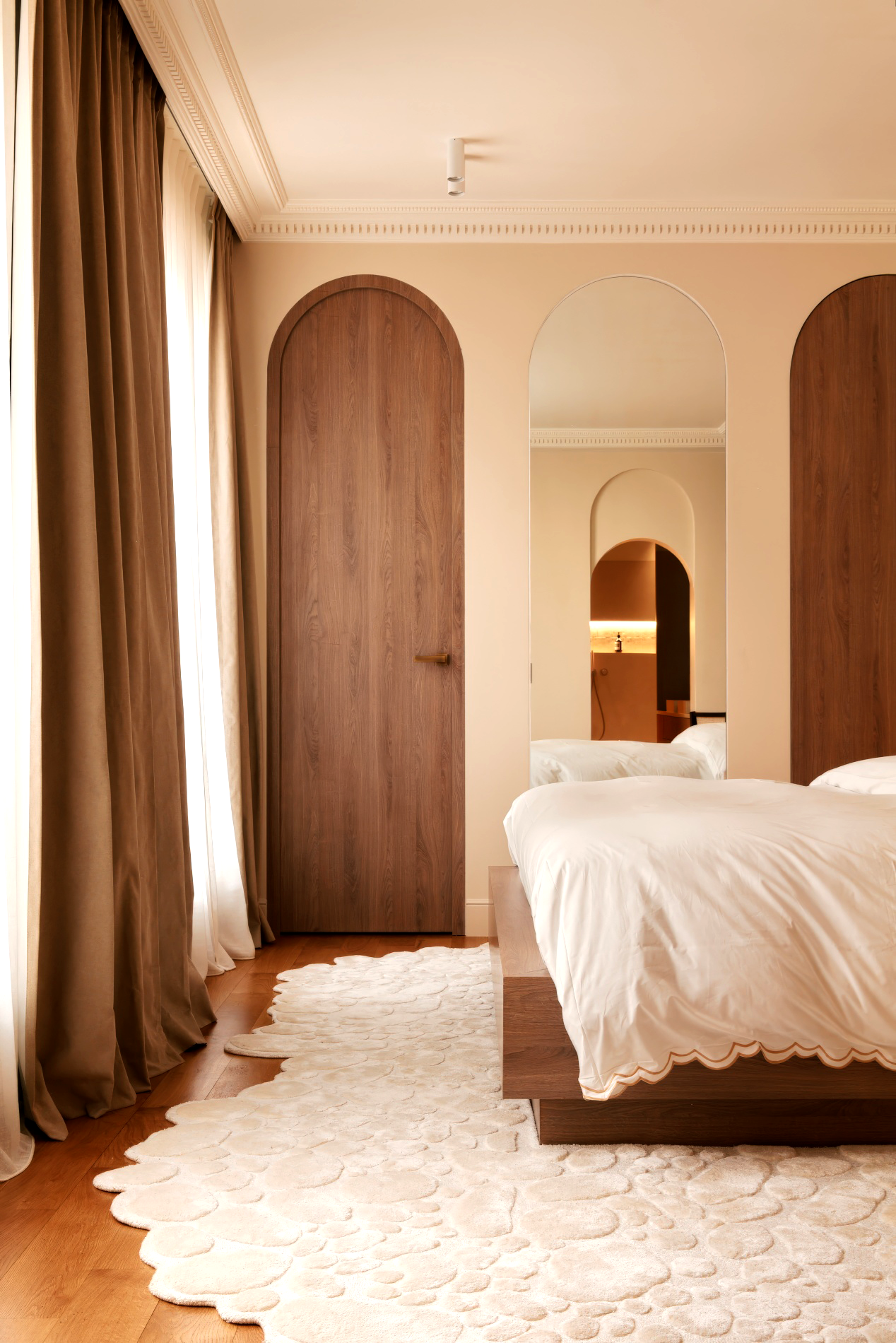 Chambre avec deux lits, tapis beige, murs crème, grand miroir encadré de bois, rideaux marron, porte en bois arrondie, décoration élégante et chaleureuse.