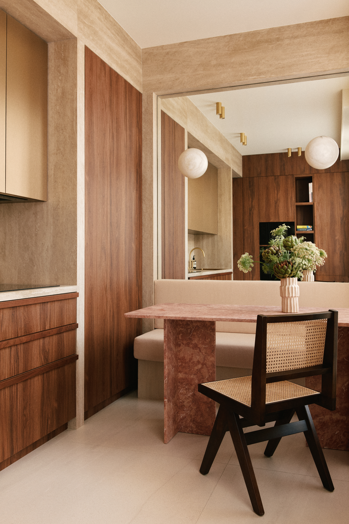 Intérieur d'une cuisine avec des murs en bois, un comptoir en marbre rose, une table assortie, un canapé beige, une chaise en bois avec assise en osier, un vase avec des fleurs, et un miroir reflétant la pièce.