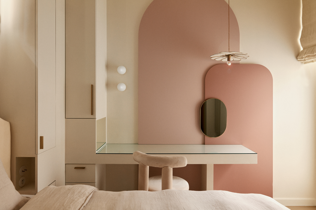 Chambre moderne avec un mur rose, un miroir ovale, un bureau blanc avec un fauteuil beige, une lampe suspendue et des armoires blanches.