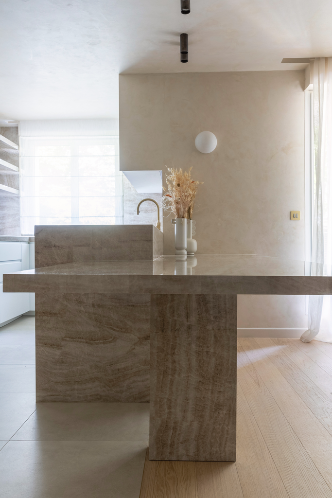 Une cuisine moderne avec un îlot en pierre beige, décorée avec un vase de fleurs séchées, près d'une fenêtre avec des rideaux blancs transparents.