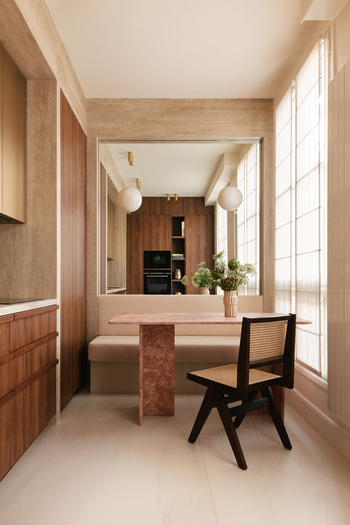 Intérieur d'une pièce lumineuse avec un coin repas, un banc rembourré beige, une table en marbre rose, une chaise en bois avec assise en osier, et de grandes fenêtres avec des rideaux blancs.