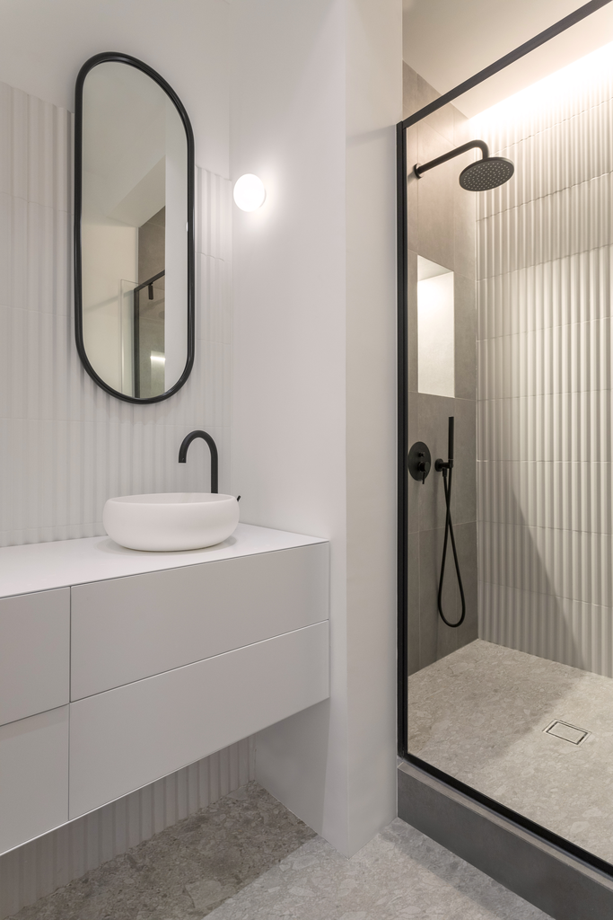 Salle de bain moderne avec lavabo, miroir oval, douche à l'italienne avec robinetterie noire, murs en carrelage blanc et gris, éclairage mural.