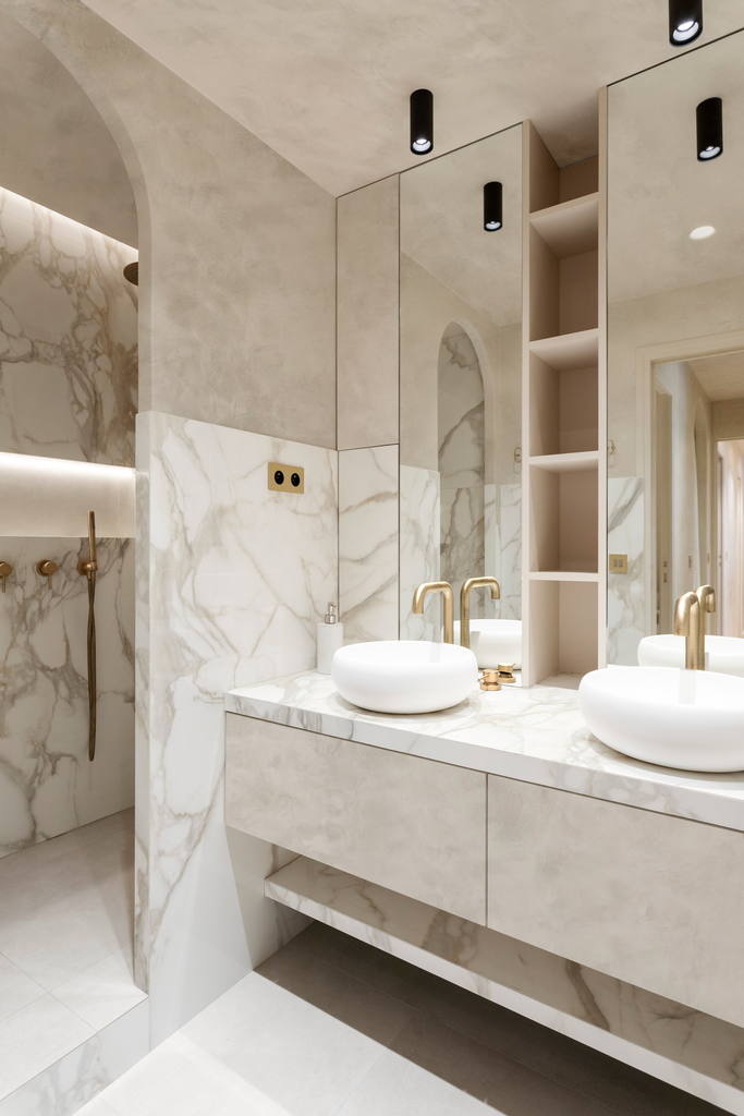 Salle de bain moderne avec un grand miroir, un double lavabo avec dessus en marbre, vasques blanches, robinets en or, et un espace douche en marbre avec des robinets dorés.