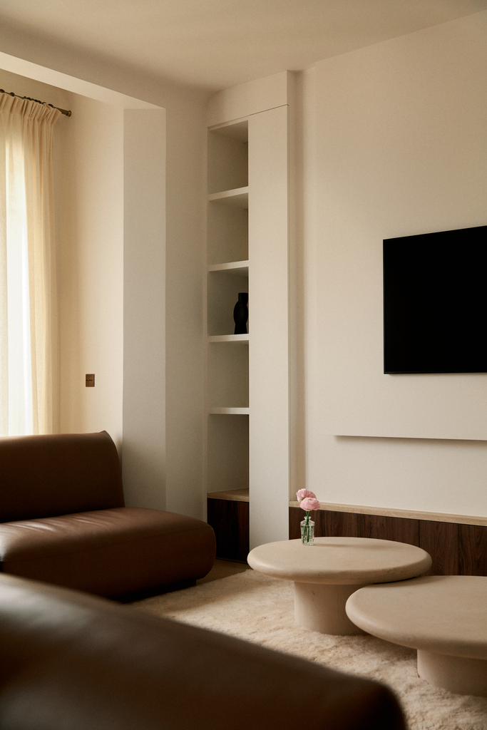 Salon avec canapé marron, deux tables basses en béton, une petite vase avec une fleur, une étagère blanche avec une décoration noire, télévision à écran plat et rideau beige.