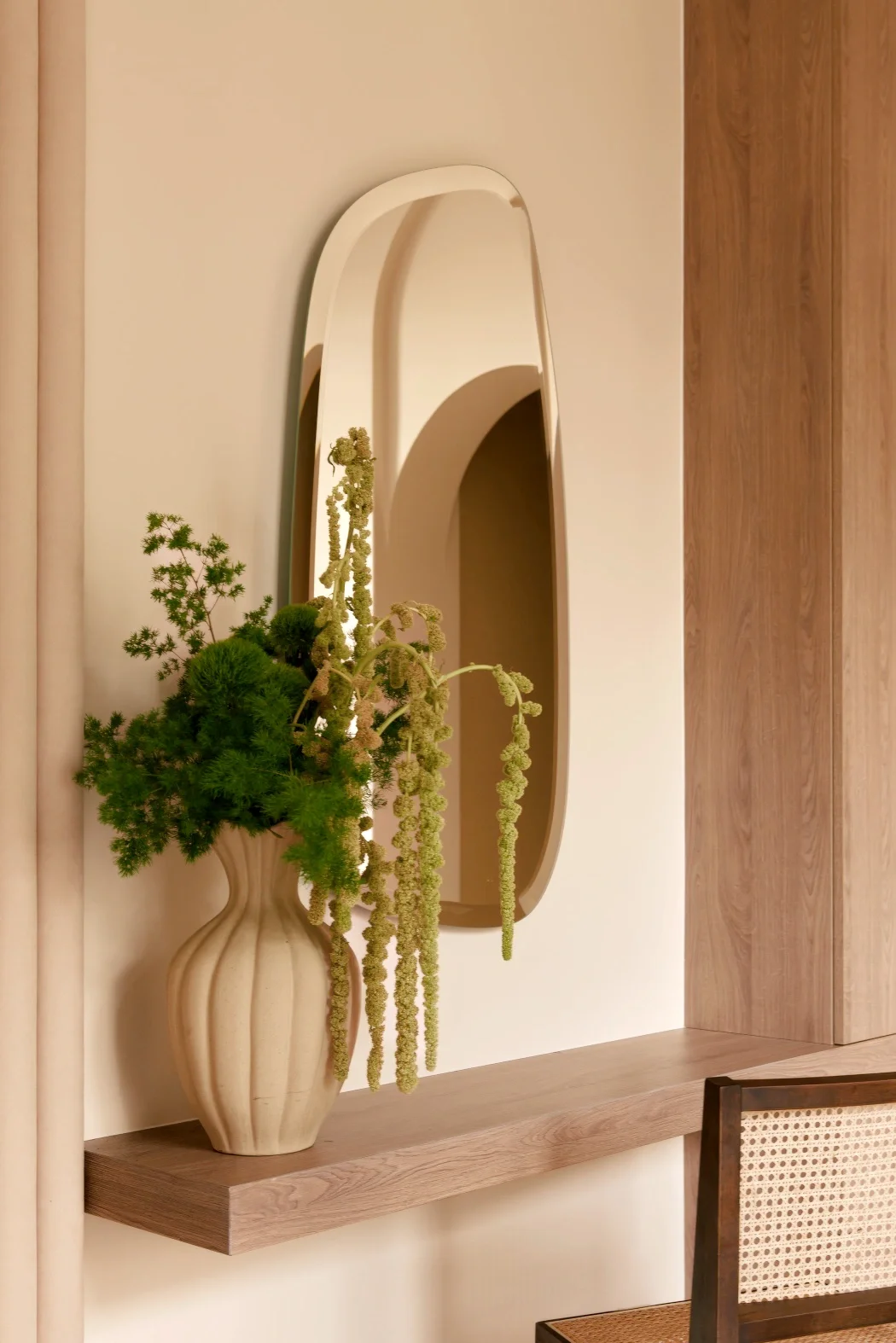 Vase en céramique beige avec des fleurs vertes et des plantes, miroir oval coklit au mur en arrière-plan, meuble en bois en dessous.
