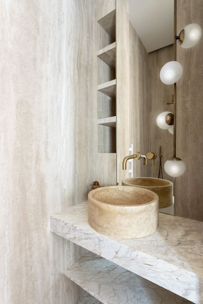 Un lavabo en pierre ronde installé sur une étagère en marbre, avec un miroir et des lampes suspendues à côté, dans une salle de bain au style moderne et minimaliste.