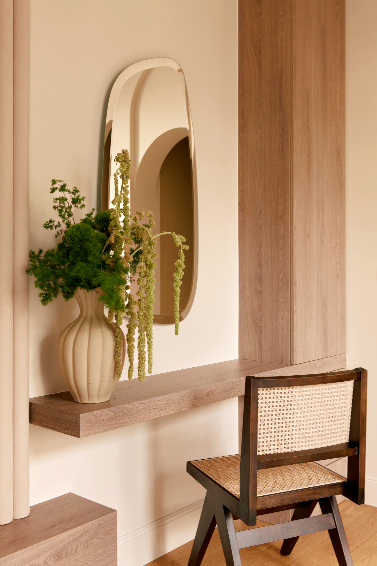 Une pièce intérieure avec une grande plante dans un vase beige, un miroir allongé, une étagère en bois clair, une table en bois et une chaise en bois avec un dossier en osier.