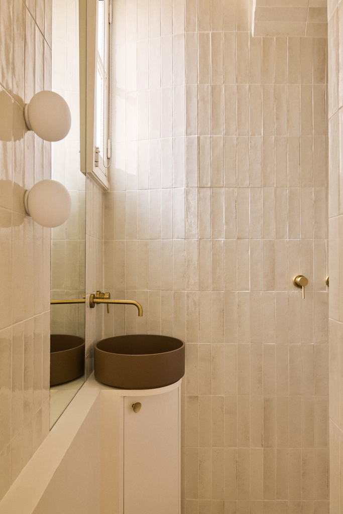 Petite salle de bain avec un lavabo rond marron, un miroir, un évier blanc sous un mur en tuiles beige clair, et un miroir mural.