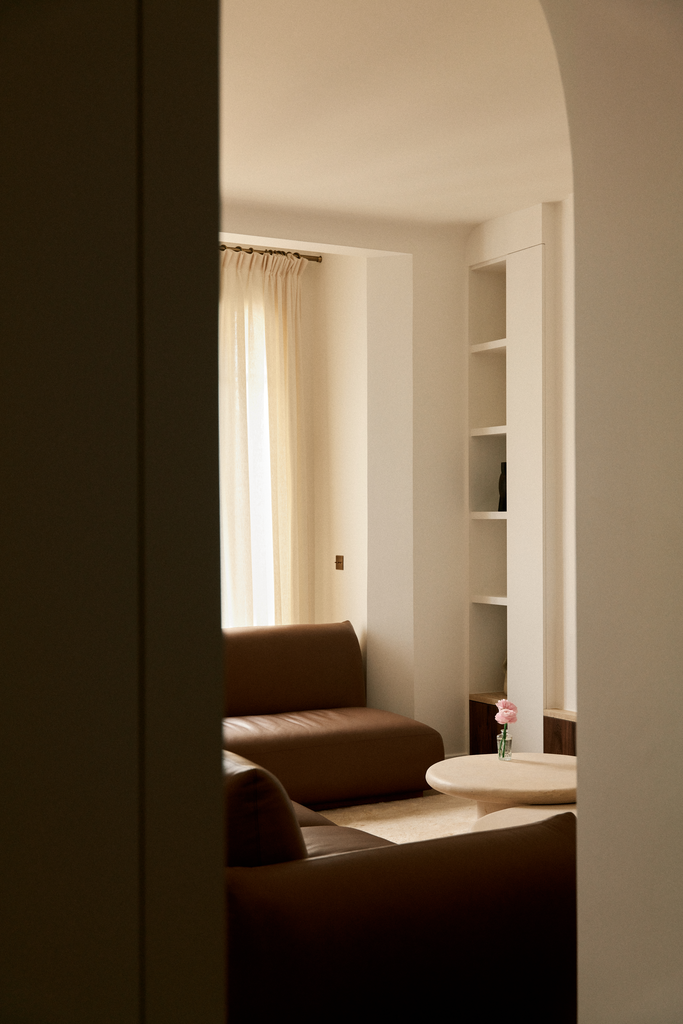Une pièce de salon éclairée par la lumière naturelle, avec un canapé marron, un petit tableau rond avec une fleur, des étagères blanches intégrées dans un mur, et un grand rideau beige sur une fenêtre.