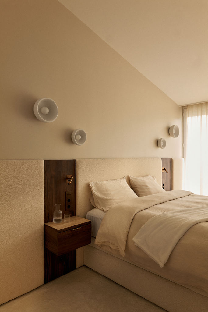 Chambre moderne avec lit double, tête de lit en blanc et brun, trois lampes murales blanches, une petite table de nuit en bois avec un verre et un flacon transparent, et un large rideau en tissu clair laissant passer la lumière.
