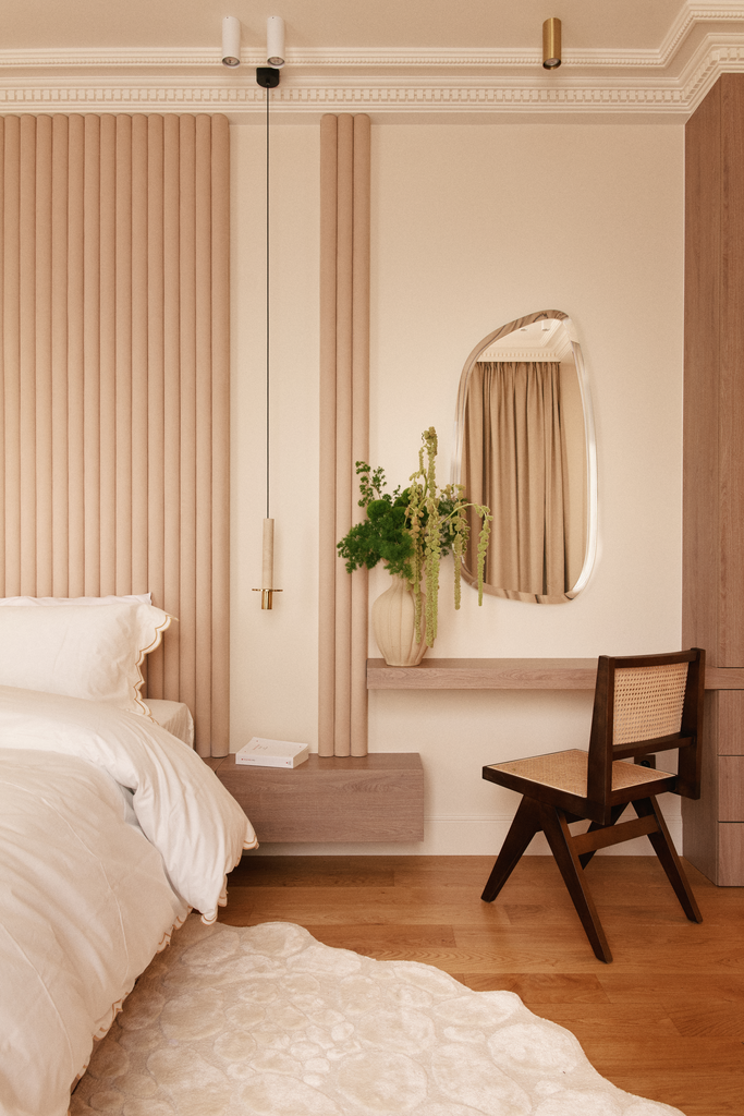 Chambre avec un lit blanc, rideaux beige, une plante dans un vase à côté d'un miroir, un fauteuil en bois, une lampe suspendue, tapis blanc sur le sol en bois.