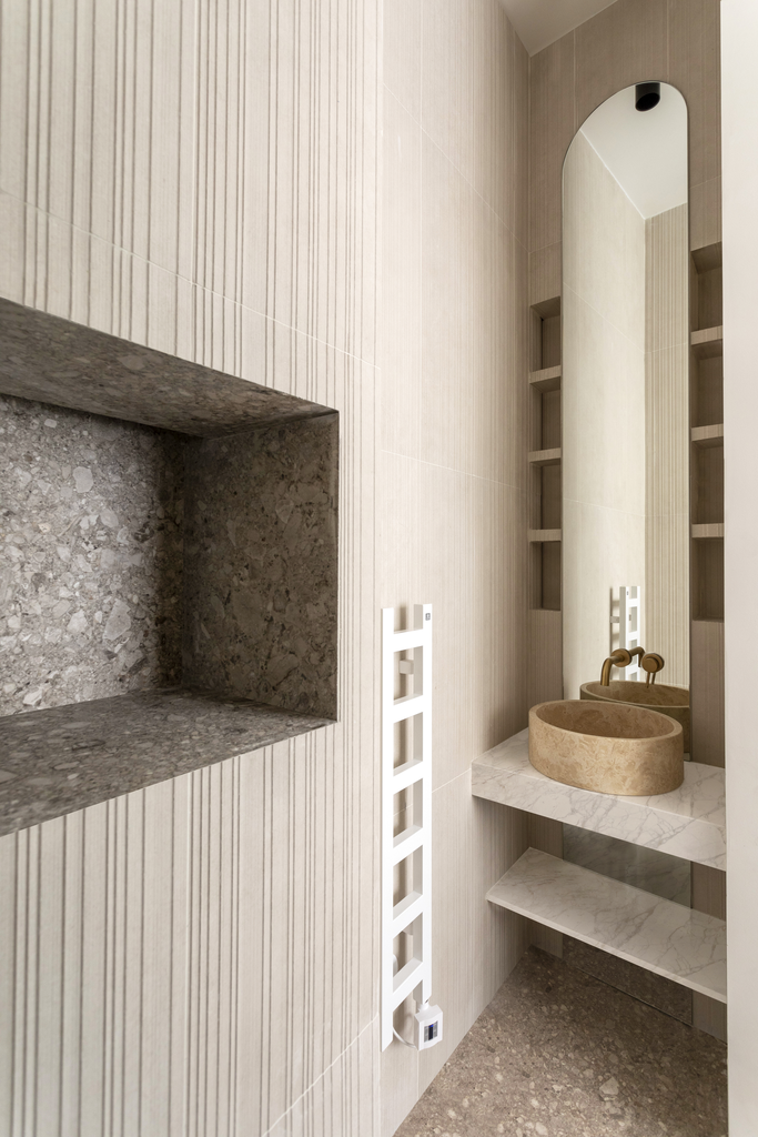 Intérieur d'une salle de bain moderne avec un lavabo en pierre beige, un miroir arqué, étagères en marbre, et une radiateur blanc vertical.