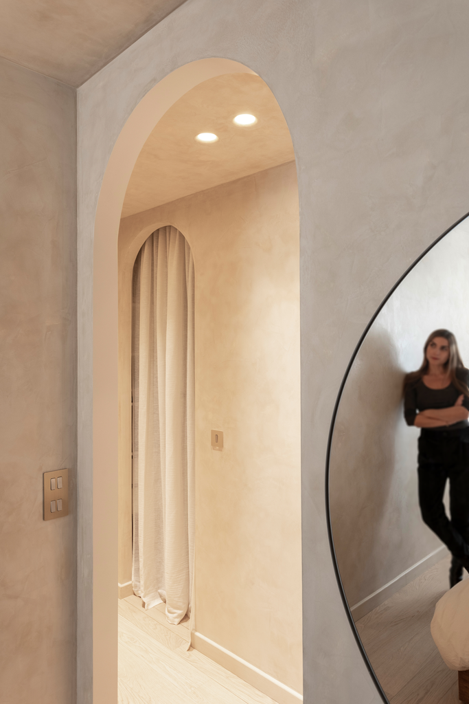 Un intérieur élégant avec un mur beige, un miroir rond, un éclairage encastré et un plaid beige devant une fenêtre avec des rideaux beige.