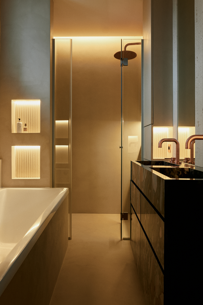 Vue intérieure d'une salle de bain moderne avec baignoire blanche, douche à l'italienne, deux lavabos avec robnets en cuivre, murs lumineux et éléments de rangement en bois.
