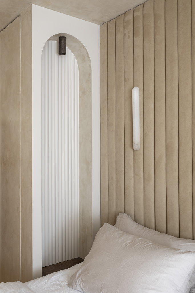 Coin de lit avec un panneau rembourré beige et une tête de lit, lumière murale moderne, mur blanc avec galerie d'art en relief noir, éléments de décoration minimalistes, ambiance chaleureuse et contemporaine.