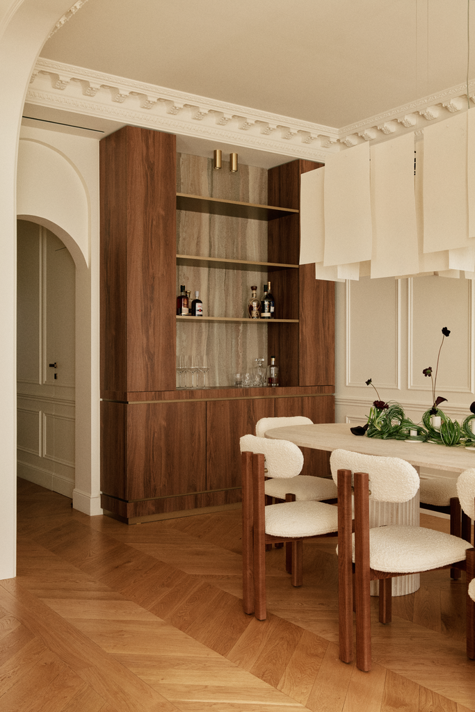 Intérieur d'une salle à manger avec une table ronde, chaises rembourrées, étagère en bois avec des bouteilles et des verres, décorations florales, et des murs ornés de moulures classiques.