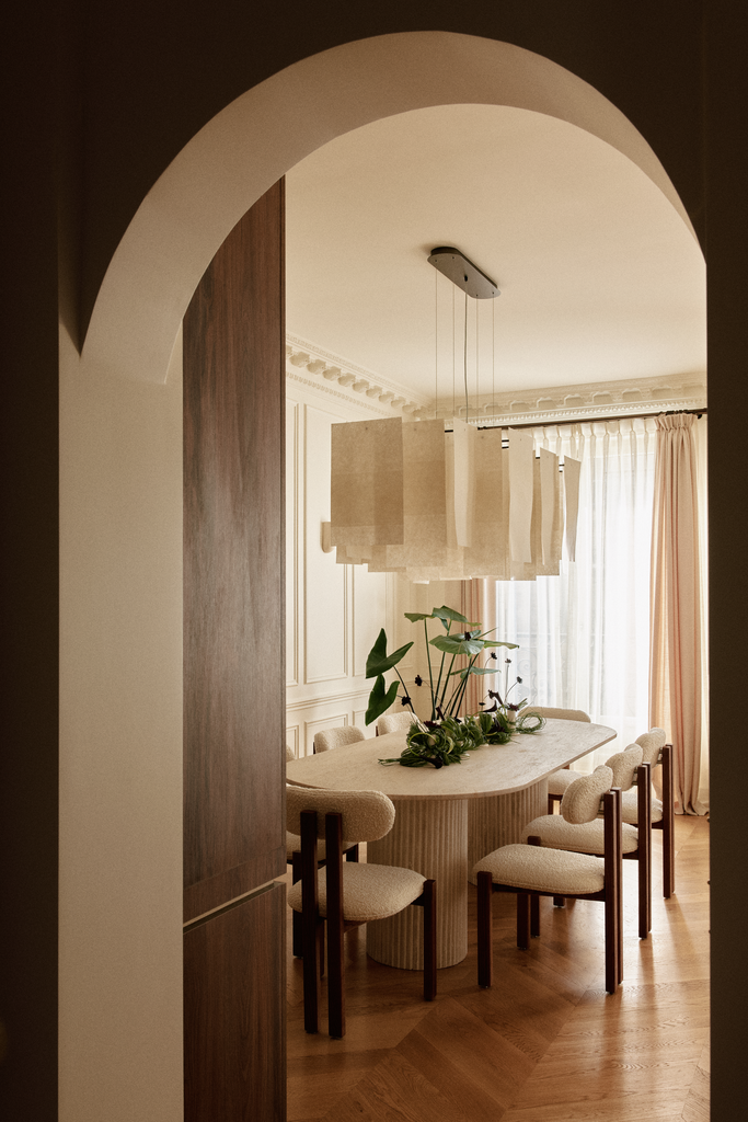 Salle à manger élégante avec une grande table ovale, six chaises rembourrées, un grand luminaire moderne, des rideaux légers laissant entrer la lumière naturelle, et une décoration florale verte au centre de la table.
