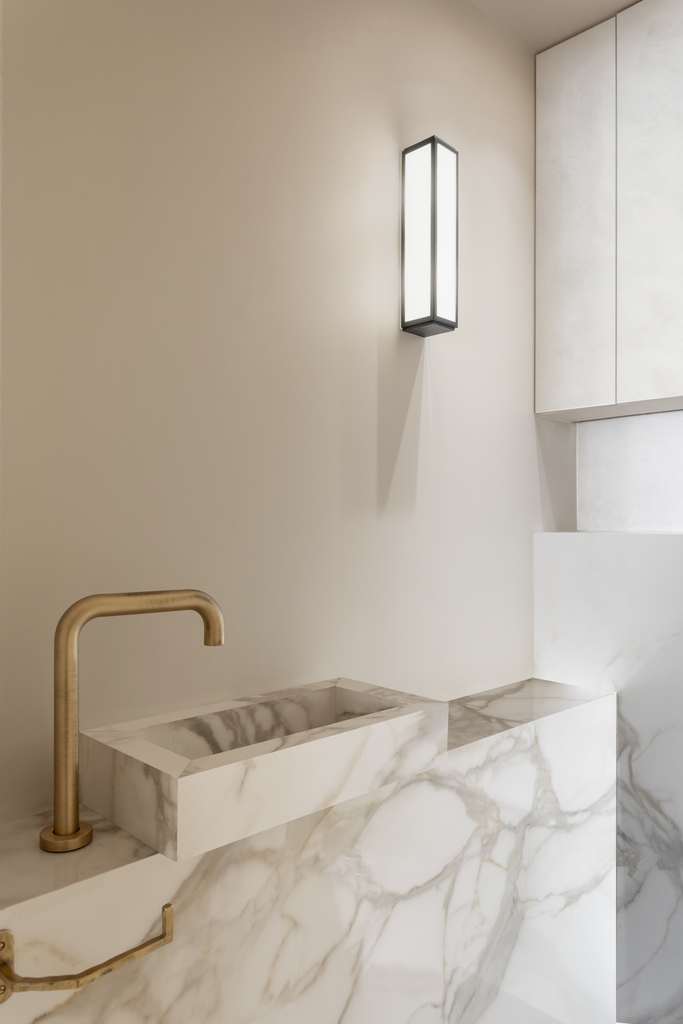 Espace de lavage ou de cuisine moderne avec un lavabo en marbre blanc et un robinet en laiton, éclairage mural rectangulaire. Murs clairs et rangement en haut.