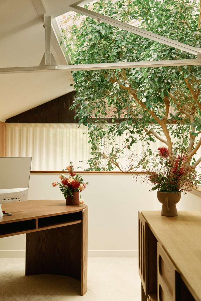 Intérieur d'une pièce avec un plafond en pente, une grande fenêtre laissant entrer la lumière, un arbre vert visible à travers la fenêtre, et deux vases de fleurs sur des meubles en bois.