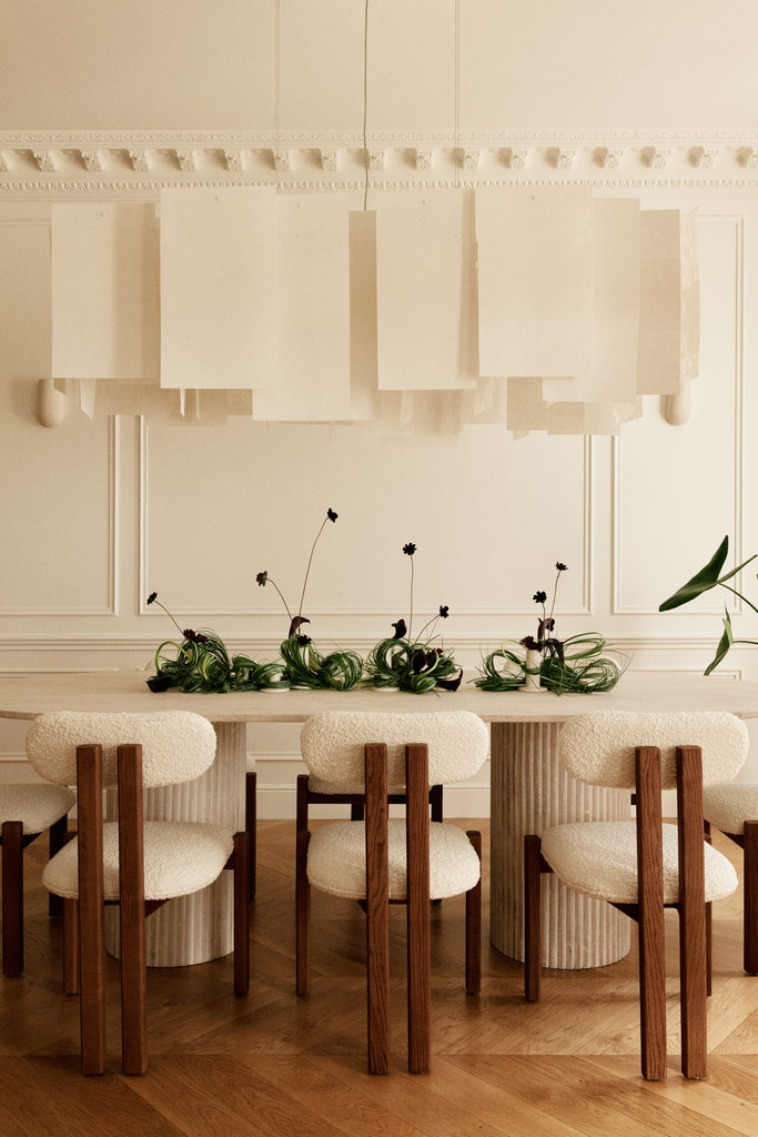 Salle à manger élégante avec une grande table en bois entourée de six chaises avec des coussins en tissu crème et des pieds en bois, décoration florale verte au centre, murs blancs avec moulures et un grand luminaire suspendu grisâtre.