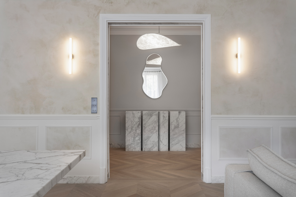 Intérieur d'une pièce moderne avec murs beige, éclairage mural, miroir abstrait, et sols en parquet. Mur de séparation en marbre blanc.