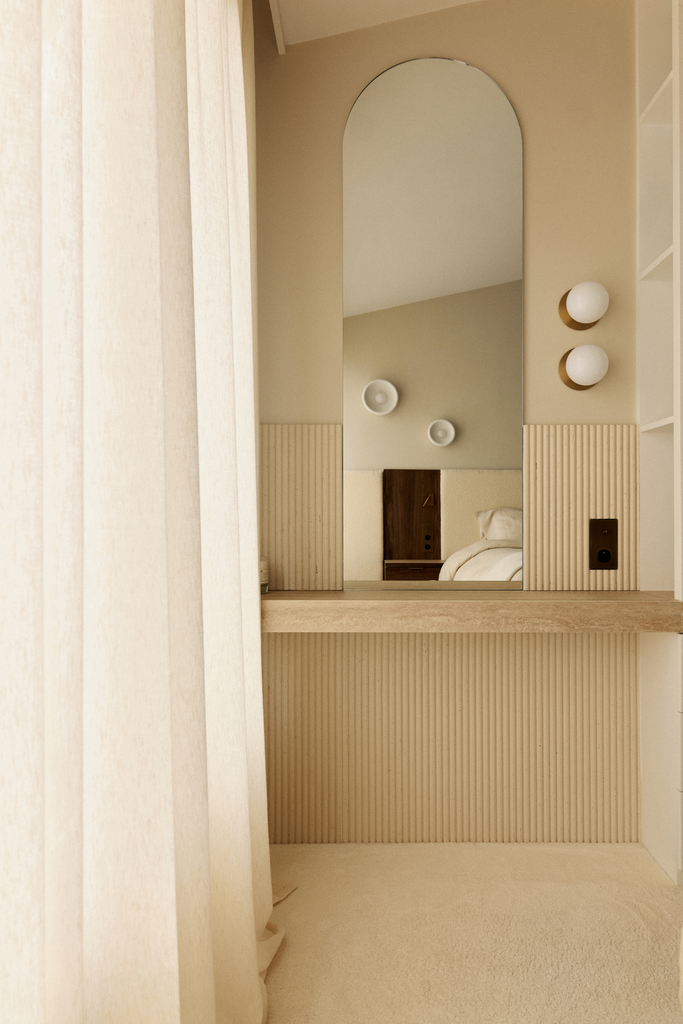 Un miroir rectangulaire avec un écho arrondi, fixé sur un mur beige, reflétant une partie d'une chambre avec un lit blanc et un meuble en bois.