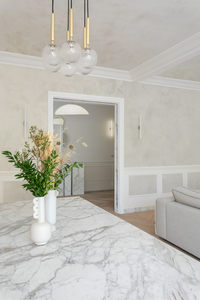 Une salle à manger avec un plafond peint en beige clair, un lustre moderne en verre et en métal, un fauteuil beige, des murs décorés avec des moulures blanches, et une table en marbre blanc avec une vase contenant des fleurs vertes et des herbes séch