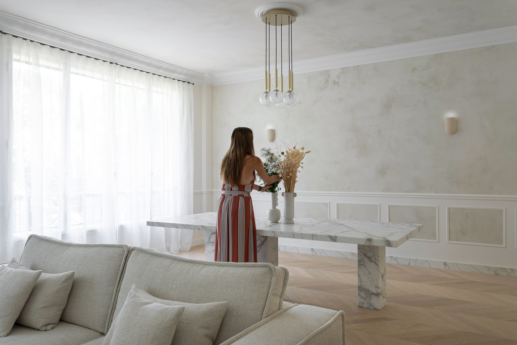 Une femme avec une robe rayée rouge, blanche et beige, debout dans un salon lumineux à côté d'une table en marbre blanc, tenant une plante. Le salon est décoré en noir et blanc avec un canapé gris, des coussins, des vases avec des fleurs séchées, et 