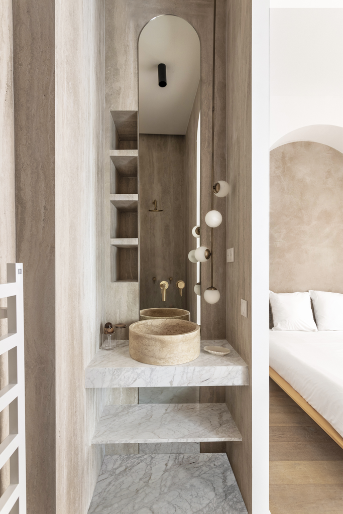 Un lavabo en pierre beige avec un miroir oval, un éclairage suspendu avec des globes blancs, et des étagères en marbre dans une salle de bain élégante et minimaliste.