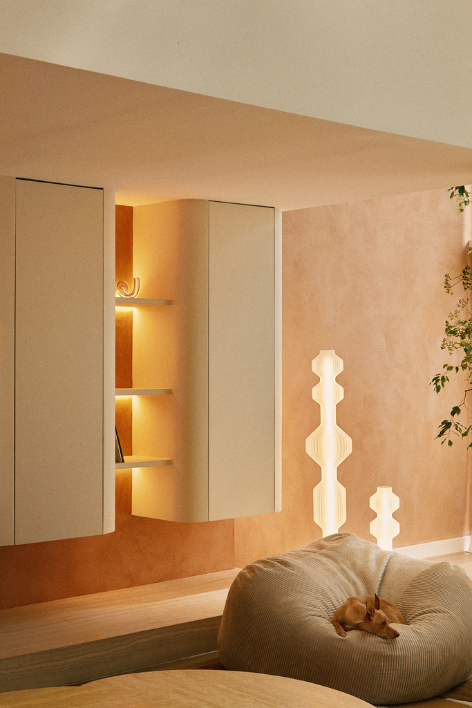 Un salon moderne avec un mur beige, une lampe décorative en forme de pyramide, une étagère intégrée éclairée, un canapé beige et un chien couché dessus.