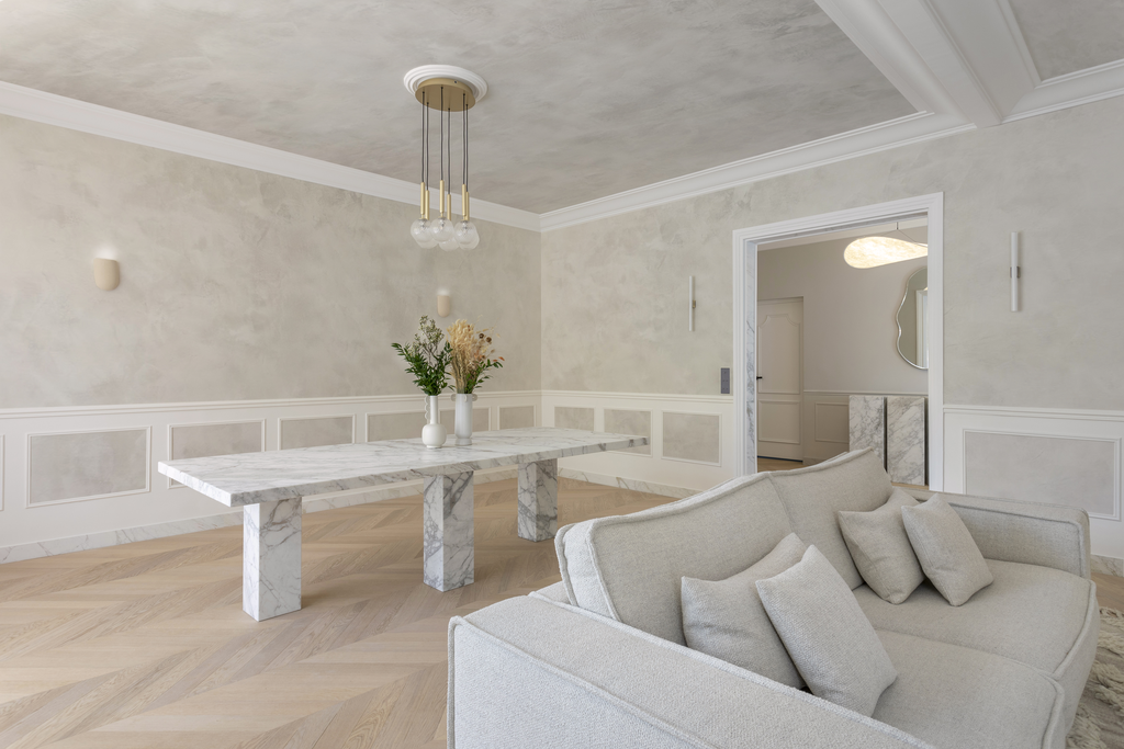 Salon moderne avec canapé beige, table en marbre, décor minimaliste, murs clairs, éclairage élégant, et porte menant à une autre pièce.