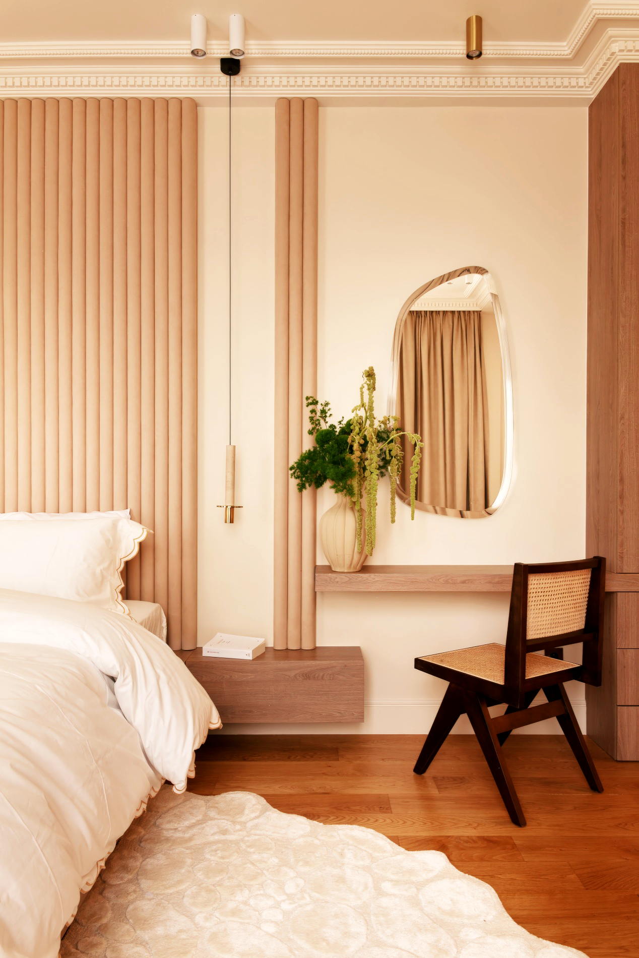 Intérieur d'une chambre avec un lit aux draps blancs, un meuble en bois, un miroir mural incurvé, un vase avec des plantes vertes, une chaise en bois avec un siège en osier, des rideaux beiges, un tapis clair et des murs crème. Décor élégant et chaleureux.